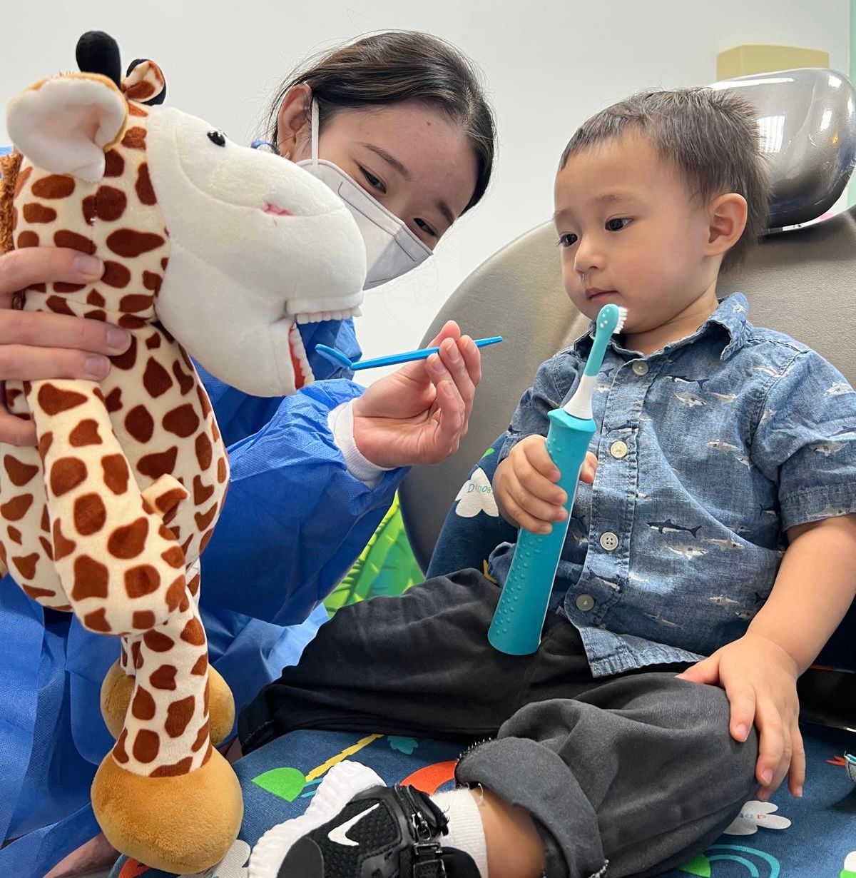 A dental hygienist uses a giraffe puppet to teach a young boy how to brush his teeth properly. The child, holding an electric toothbrush, watches the demonstration closely.